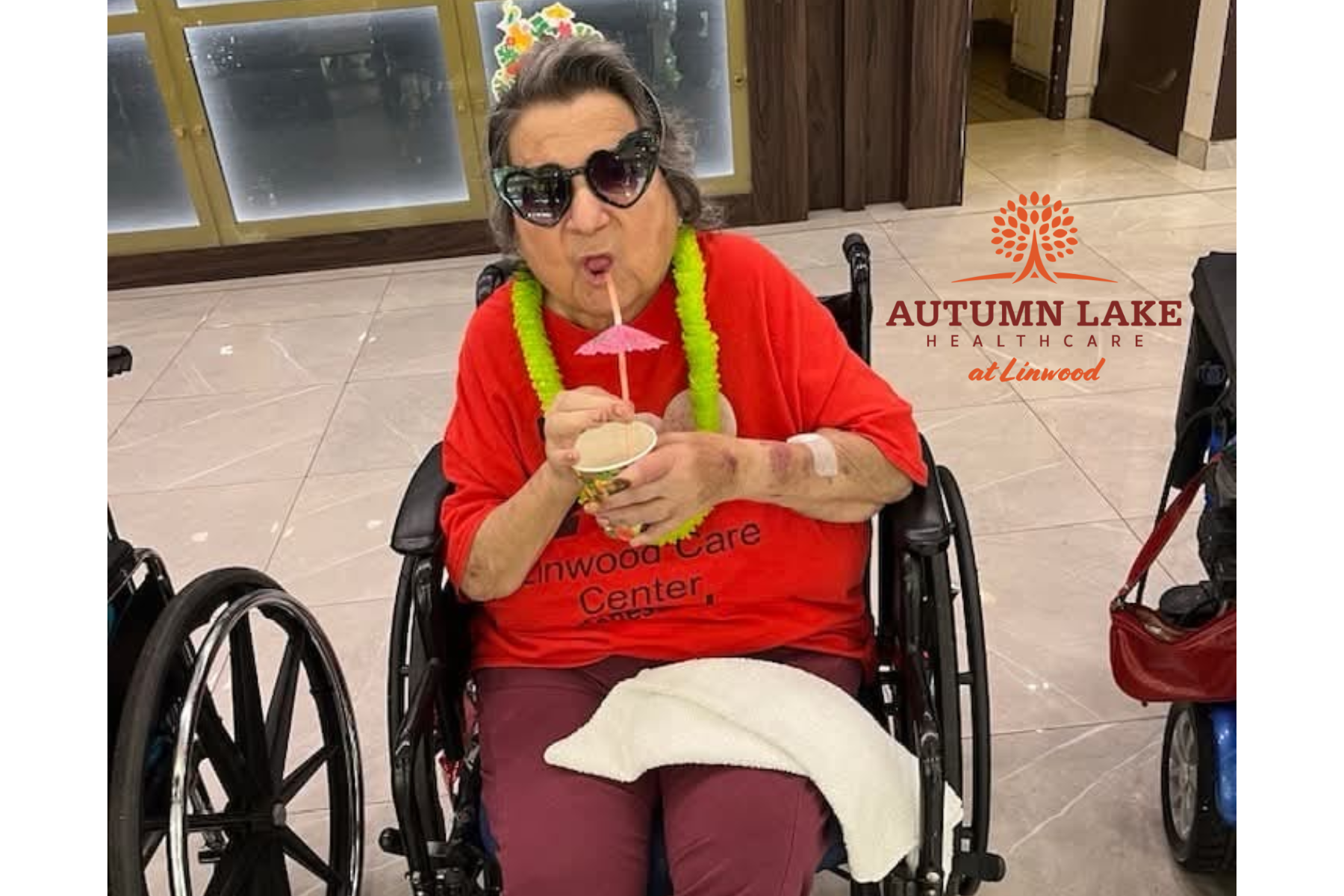 A nursing home resident in a wheelchair wears heart sunglasses and a lei while sipping a festive drink.