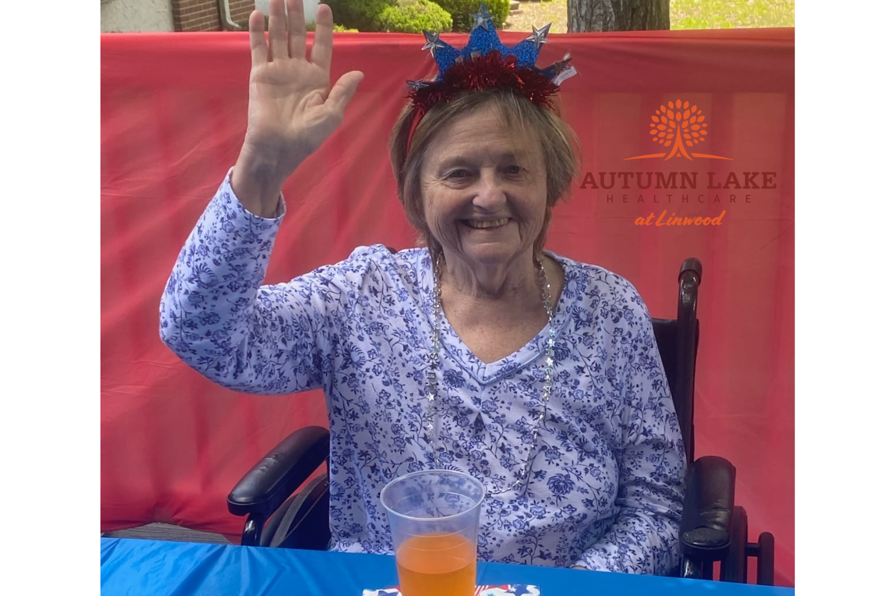 A smiling resident in a wheelchair waves at a patriotic-themed event at a rehabilitation care center.