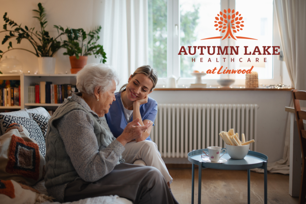 A nurse in blue scrubs smiling at a senior woman seated in a cozy living area.