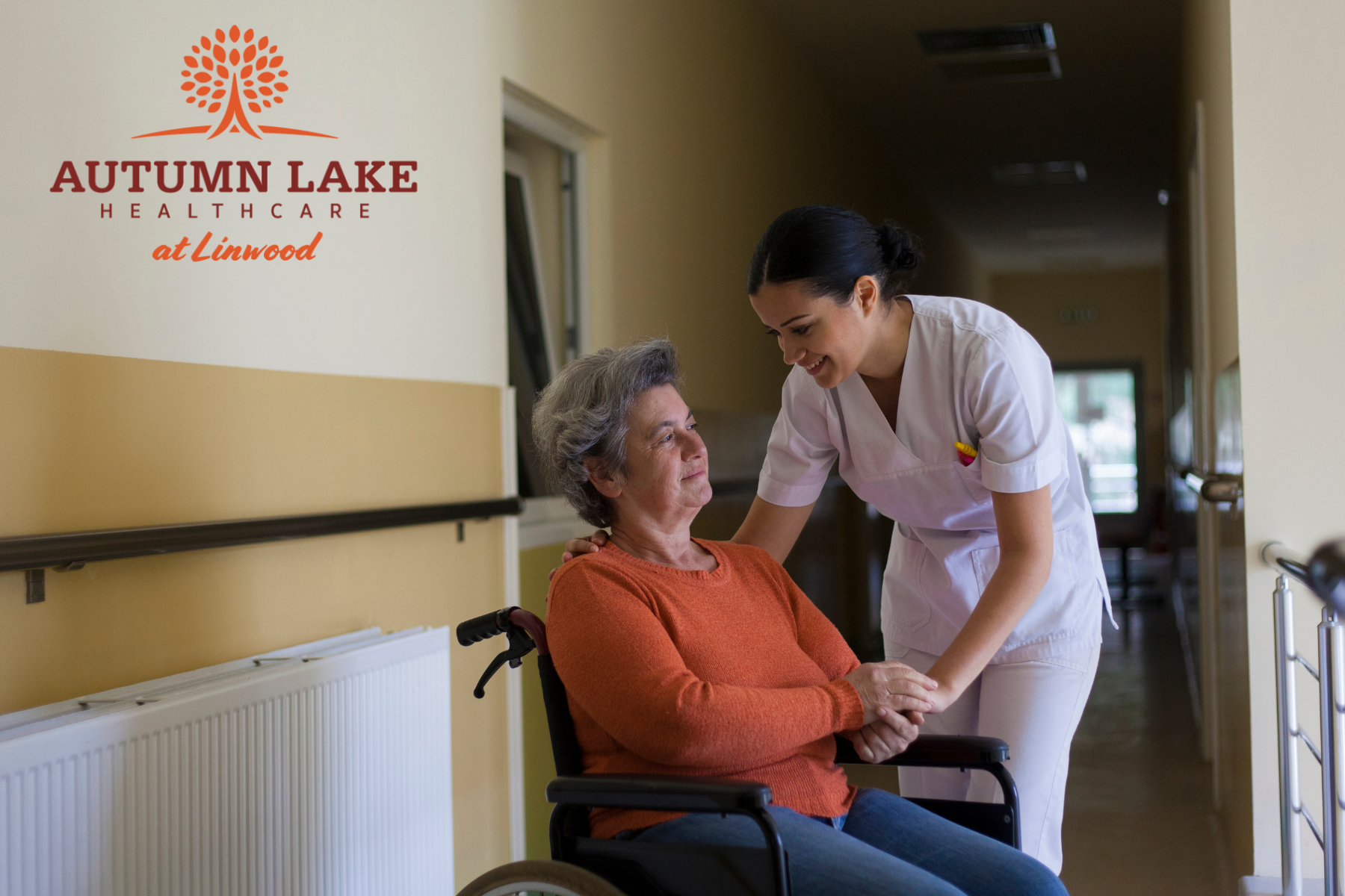 A nurse in white scrubs comforts a senior resident in a wheelchair in a hallway at Autumn Lake Healthcare at Linwood.
